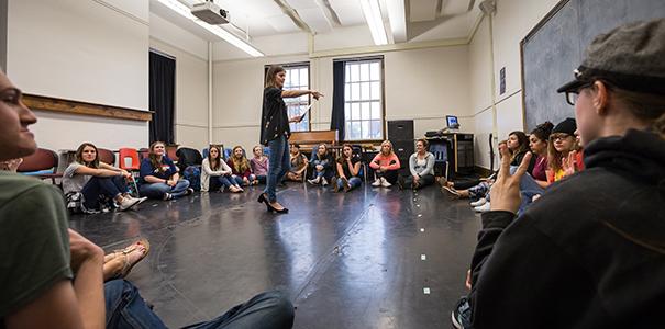 instructor standing in circle of students in a large room conducting acting class